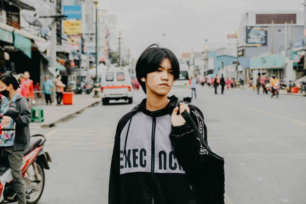 A young person with a jacket stands confidently in an urban street environment.